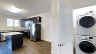 Located at Windsor Castle, this single-floor space offers a thoughtfully designed interior that blends modern aesthetics with functional living. The kitchen is a standout feature, showcasing rich dark cabinetry that extends to the upper walls, providing ample storage. Stainless steel appliances, including a full-size refrigerator, gas range, and built-in microwave, lend a contemporary feel to the space. Light-toned countertops contrast elegantly with the dark cabinetry, creating a visually balanced cooking area. The flooring throughout features wood-style planks in a warm, medium tone, adding warmth and cohesion to the interior. Crisp white walls and white baseboards keep the space feeling bright and open, complementing the overall modern design. A louvered interior door with a brushed nickel handle adds a subtle architectural detail, contributing to the clean, well-finished look of the space. The open layout between the kitchen and adjoining areas promotes an easy flow, making the space feel welcoming and practical for everyday use.