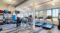 This fitness center offers a spacious, single-floor layout thoughtfully designed to accommodate a full range of workout needs. The interior features high ceilings with exposed structural beams and sleek linear pendant lighting, contributing to an open, energizing atmosphere. Large, multi-pane windows line the walls, flooding the space with natural light and offering views of the surrounding greenery.

The flooring throughout consists of stylish modular carpet tiles arranged in a geometric pattern of neutral tones with subtle yellow-green accents, providing both durability and visual interest. Sturdy white structural columns are positioned throughout the floor, defining zones without obstructing flow.

The cardio area is equipped with multiple treadmills, ellipticals, and stationary bikes, all positioned to face the windows. The strength training zone includes a full rack of dumbbells displayed along a wall-mounted mirror, various cable machines, a power rack with an Olympic barbell and bench, and a dedicated functional trainer. Exercise balls are neatly stored on a wall-mounted rack, and flat-screen TVs are mounted at key viewing angles throughout. An adjacent ancillary room provides additional storage and utility space. The overall ambiance is clean, modern, and professional, making this fitness center a highly functional and welcoming environment for exercise.