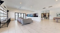 Located at 1614 Hill St in Grand Prairie, Texas, this contemporary single-floor community amenity space offers a generously sized, open-plan layout designed for both relaxation and social gatherings. The interior is anchored by large-format light tile flooring that runs seamlessly throughout, complemented by crisp white walls and recessed lighting that create a bright, airy atmosphere.

The lounge area features multiple plush seating arrangements, including a large sectional sofa and accent chairs arranged on bold black-and-white striped area rugs, centered around a wall-mounted flat-screen television. Expansive black-framed glass doors and industrial-style roll-up windows flood the space with natural light and provide direct access to an outdoor balcony with views of the surrounding community.

A standout dining and kitchen zone showcases a substantial wood communal table surrounded by powder-blue upholstered chairs, illuminated by a series of elegant amber pendant lights. The kitchen behind features stainless steel appliances, built-in wall ovens, and a striking arched architectural niche. A game area with a pool table and bistro-style high-top seating adds a recreational dimension. A glass-partitioned private room offers a more intimate setting for smaller gatherings, rounding out this well-appointed, versatile space.