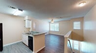 This 3-bedroom, 2-bathroom home offers a practical and comfortable layout designed to maximize everyday living. The open-concept kitchen and living area form the heart of the home, where wood-look flooring flows seamlessly underfoot. The kitchen is equipped with white cabinetry, granite-look countertops, a peninsula with a built-in sink, and black appliances including a refrigerator, range, and dishwasher — all beneath a textured ceiling with overhead lighting. The adjoining living room is bright and airy, featuring two windows with blinds and a ceiling fan with light kit, providing both ventilation and natural light. A carpeted staircase with a wooden handrail leads to the upper level entry area, where a windowed landing offers additional natural light. Each of the three bedrooms features plush wall-to-wall carpeting, a window with blinds, and ceiling-mounted lighting, with one bedroom also including a ceiling fan. The two bathrooms are clean and functional; one features a granite-look vanity countertop with white cabinetry and a large mirror, while the other includes a full tub-shower combo with tile surround and wood-look flooring. Throughout the home, neutral wall tones and consistent finishes create a cohesive and inviting atmosphere.