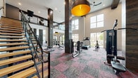 This two-level fitness center offers a generously proportioned workout environment spread across a larger main floor and a secondary upper level. The ground floor immediately impresses with its soaring double-height ceilings, dark wood-clad structural columns, and exposed beam detailing that lend a refined, contemporary aesthetic. Large windows along the perimeter flood the space with natural light while providing views of the surrounding outdoor areas. Bold pendant lighting with gold-toned interiors adds a striking visual contrast against the crisp white walls. Patterned carpet tiles in muted red and grey tones define the strength and cardio zones, where a full selection of cable machines, free weights, benches, and specialty equipment is thoughtfully arranged. A functional training station includes suspension trainers, medicine balls, kettlebells, and plyo boxes. A glass-railed staircase with wooden treads connects to the upper level, which features striped rubberized flooring and houses treadmills, ellipticals, and stationary bikes alongside a dedicated open studio space equipped with a wall-mounted display screen, stability balls, and additional free weights—ideal for group fitness or individual training sessions.