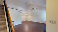 This multi-story home spans three floors and showcases a thoughtfully designed interior that balances open space with functional living. The main living area immediately impresses with its dramatic vaulted ceilings that rise to a striking height, creating a grand sense of volume and airiness. A ceiling fan with an extended drop rod is centrally positioned to keep the space comfortable year-round, while a pendant chandelier adds a touch of elegance and provides ambient lighting. The flooring throughout features rich, dark hardwood-style planks that lend warmth and a polished, cohesive aesthetic to the space. Crisp white walls and clean lines contribute to a bright, neutral palette that enhances the sense of openness. A partial wall divider subtly separates living zones while maintaining the flow of light and sight lines across the open floor plan. Visible HVAC vents are integrated into the ceiling and walls, reflecting a practical approach to climate control. The staircase, partially visible, connects the floors with classic wooden railings, adding a traditional architectural touch. Overall, this home offers a welcoming and spacious atmosphere, with standout architectural features that make it both visually impressive and highly livable across all three levels.