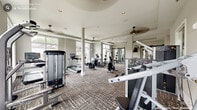 This fitness center is thoughtfully designed across a single floor, offering two interconnected workout zones that together provide a comprehensive exercise environment. The main cardio and strength area features tray ceilings with ceiling fans, structural columns, and an abundance of natural light streaming through large floor-to-ceiling windows that frame views of the surrounding community. The neutral gray walls and patterned carpet flooring create a clean, modern aesthetic throughout. This primary zone is outfitted with multiple treadmills, ellipticals, stationary bikes, and a selection of selectorized strength machines, along with a wall-mounted flat-screen TV and full-length mirrors that enhance the sense of space.

A secondary room, accessible through stylish black-framed barn-style glass doors, offers a dedicated functional training space finished with tile-pattern flooring and a tray ceiling. This area houses a multi-use cable machine, rowing machine, dumbbell rack, resistance bands, and exercise balls, making it ideal for versatile, focused workouts. A wall of mirrors amplifies the room and provides visual feedback during training sessions. Together, the two zones create a well-rounded fitness facility that balances cardio, strength, and functional training in a bright, modern, and inviting setting.
