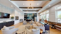 Located at 101 McCaw St in Mauldin, South Carolina, this single-floor community clubroom offers a thoughtfully designed open-concept layout that seamlessly blends comfort and functionality. The main lounge area is anchored by a tray ceiling adorned with dark wood beams and a mid-century modern splay chandelier, creating an inviting focal point above the generous arrangement of neutral-toned sofas and accent chairs. Light hardwood flooring runs throughout, contributing to the warm, airy ambiance of the space.

The kitchenette is a standout feature, showcasing dark cabinetry with gold hardware, a striking teal herringbone tile backsplash, stainless steel appliances including a French door refrigerator, open shelving, and a sleek white countertop with an undermount sink — all topped by a statement pendant light.

A separate dining and game zone is bathed in natural light from floor-to-ceiling windows and glass doors that open directly to an outdoor pool area, offering scenic views and easy access to outdoor amenities. This section also features a shuffleboard table, round dining tables, and upholstered seating.

Board and batten wainscoting lines multiple walls, adding architectural interest, while recessed lighting throughout ensures a bright, welcoming atmosphere suitable for both casual lounging and community events.