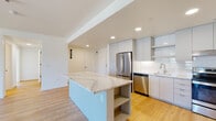 This single-floor residence is designed with a clean, contemporary aesthetic that emphasizes open living and functional design. The kitchen serves as the centerpiece of the main living area, featuring flat-front cabinetry in a soft grey-white finish, a white subway tile backsplash, and a full suite of stainless steel appliances including a French door refrigerator, electric range, and dishwasher. A large kitchen island with striking veined marble-look countertop provides additional prep space and open shelving beneath, seamlessly connecting to the expansive living area.

Warm, light-toned hardwood flooring runs throughout the entire residence, lending a cohesive and inviting feel to each room. The living area is bright and airy, benefiting from large windows that allow natural light to pour in while offering views of the surrounding neighborhood. Recessed lighting supplements natural light throughout.

The bedroom includes a generously sized walk-in closet outfitted with adjustable wire shelving and double hanging rods. The bathroom is finished in crisp white tones, with an undermount sink set into a solid white countertop, a large wall mirror, and a glass-enclosed shower. A stacked washer and dryer unit is conveniently housed within a dedicated laundry closet.