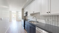 This thoughtfully designed 2-bedroom, 1-bathroom unit offers a cohesive and modern living environment across a single floor. The open layout is anchored by a bright living room featuring warm hardwood-style flooring, large windows, and a glazed exterior door that floods the space with natural light and provides outdoor views of surrounding greenery. The neutral color palette of soft whites and greiges creates a clean, inviting ambiance throughout.

The galley-style kitchen is a standout feature, showcasing a striking two-tone cabinet design with navy blue lower cabinets and crisp white upper cabinets, complemented by dark stone countertops and a vertically laid white tile backsplash. Stainless steel appliances, including a range, microwave, and dishwasher, complete this well-appointed kitchen.

Both bedrooms offer a calm and functional retreat, with the same warm hardwood-style flooring and neutral wall tones continuing throughout. The primary bedroom provides a comfortable amount of space for furnishing.

The bathroom is finished with classic black-and-white hexagonal mosaic floor tiles, large-format white wall tiles surrounding the tub-shower combination, and a glass shower door, delivering a clean and timeless aesthetic. A central hallway connects all rooms efficiently, completing this practical and stylish unit.