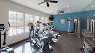 This fitness center offers a comprehensive workout environment designed to support a wide range of exercise routines. The space is organized into distinct functional zones, providing both cardio and strength training areas within a single, open floor plan. Durable rubber flooring covers the entire space, providing both safety and comfort during workouts.

The cardio zone is well-stocked with multiple treadmills, elliptical trainers, and a stationary recumbent bike, several of which are Precor brand machines. Large windows along the walls flood this area with natural light and offer views of an outdoor pool area with yellow umbrellas, creating an inviting and energizing atmosphere. Ceiling fans mounted overhead help maintain air circulation throughout. A wall-mounted flat-screen television is positioned for easy viewing during cardio sessions, accompanied by a small shelf below.

The strength training area features a multi-functional cable machine surrounded by stability balls and is set against a bold teal accent wall, adding a vibrant visual contrast to the otherwise neutral color palette of white and gray. A large mirror spans one wall, enhancing the sense of space and allowing for form checks during exercise. Framed informational posters and fitness guides are displayed throughout, adding a practical, health-focused touch to the overall environment.