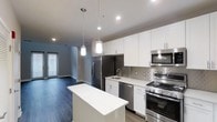 This 2-floor space showcases a contemporary design with a well-appointed kitchen as its centerpiece. The kitchen features crisp white shaker-style cabinetry extending to the ceiling, offering generous storage throughout. A suite of stainless steel appliances includes a French door refrigerator, gas range, built-in microwave, and dishwasher, all complementing the clean, modern aesthetic. A decorative geometric tile backsplash adds visual interest behind the cooking area, while a undermount sink with a sleek faucet is positioned along the perimeter counter. A central island with a bright white countertop provides additional workspace and casual seating potential. Two pendant lights hang above the island, adding warm, focused lighting to the space. Dark hardwood-style flooring runs throughout, creating a striking contrast against the white cabinetry and light countertops. Recessed lighting supplements the pendant fixtures, ensuring the space is well-lit. The open-concept layout connects the kitchen to the living area beyond, where natural light filters in through a window, enhancing the overall sense of openness across both levels.