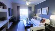 This single-floor space centers around a well-appointed living room that combines comfort with contemporary style. The room is defined by a striking dark accent wall, which serves as a bold backdrop for framed wall art and provides a strong visual contrast against the lighter surrounding walls trimmed with classic crown molding. A ceiling fan with a built-in light fixture is centrally positioned overhead, contributing both to the room's functionality and its finished aesthetic.

Natural light fills the space through large sliding glass doors dressed with sheer white curtains and dark draping panels, offering an effortless transition to an outdoor balcony with railing. The seating area is anchored by a plush white sofa accented with patterned throw pillows in navy, rust, and neutral tones, complemented by additional seating and a sleek glass-top coffee table resting on a woven area rug. A media console along the opposite wall accommodates a flat-screen television, while lush indoor plants add organic warmth to the overall composition. The space presents a cohesive and inviting atmosphere suited for everyday living and entertaining.