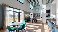 This single-floor amenity lounge is a thoughtfully designed communal space that seamlessly blends social and recreational functions. The most striking architectural feature is the soaring double-height ceiling in the main lounge area, accentuated by teal-painted exposed beams and a clerestory row of windows that flood the interior with abundant natural light. Large glass doors open toward an outdoor terrace, offering city skyline views and reinforcing the connection between indoor and outdoor spaces.

The herringbone-patterned wood-look tile flooring unifies the main gathering areas, which are furnished with teal upholstered chairs, a comfortable gray sectional sofa, accent armchairs, and a large wall-mounted television. An industrial-style open bookshelf anchors one corner, complemented by warm brass sconce lighting. A foosball table adds a playful recreational element to the space.

A dedicated wet bar area features a marble-topped island with bar seating, brass pendant lighting, open shelving with glassware, a mounted television, and a beverage refrigerator. An adjacent window-side counter with mixed-tone seating provides a casual workspace or dining area. The overall ambiance is sophisticated yet approachable, combining contemporary design with inviting, comfortable furnishings.