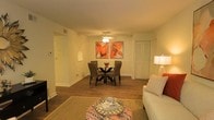 This single-floor residence showcases an open-concept layout that seamlessly connects the living and dining areas into one cohesive, functional space. The flooring throughout is rich hardwood, lending warmth and continuity to the interior. A ceiling fan with an integrated light fixture is centrally positioned, providing both airflow and ambient illumination to the combined living-dining zone.

The dining area is anchored by a round glass-top table paired with woven rattan-style chairs, offering a casual yet stylish setting. Above the dining table, two bold, orange-toned abstract artworks serve as a striking focal point against the neutral, light-colored walls. A small decorative niche in the wall adds an architectural detail that breaks the uniformity of the surfaces.

The living area accommodates comfortable seating, with a sofa accented by patterned throw pillows. A round, upholstered ottoman with a decorative tray sits atop a textured area rug, grounding the seating arrangement. A floor lamp provides additional warm lighting, contributing to the cozy, inviting ambiance of the space. A closet with a paneled door is visible, offering practical storage within the main living area.