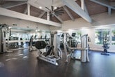 Located at 888 Davis St in San Leandro, CA, this fitness center offers a thoughtfully designed workout environment that balances functionality with architectural character. The space is defined by its impressive vaulted ceiling, showcasing exposed wood planking and bold white structural beams that soar overhead, creating an open and energizing atmosphere. A large ceiling fan anchors the apex of the ceiling, contributing to air circulation throughout the room.

The flooring consists of durable rubber-textured material, providing traction and impact absorption suitable for a range of workout activities. Multiple windows along the walls flood the interior with natural light and offer pleasant views of the surrounding outdoor greenery and pool area, enhancing the overall workout experience.

The fitness center is comprehensively equipped for both cardio and strength training. The cardio section features a row of treadmills and stationary bikes — including upright and recumbent models — all positioned in front of a mirrored wall that amplifies the sense of space. The strength training area houses Hoist-branded cable and resistance machines, a dumbbell and kettlebell rack, weight benches, and an assortment of stability and medicine balls. Glass double doors provide easy access to the exterior, further connecting the indoor space to its outdoor surroundings.