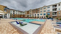 This impressive outdoor amenity space centers around a large, resort-style swimming pool complex designed for both relaxation and recreation. The pool area is thoughtfully divided into distinct zones: a spacious main lap-style pool with vivid turquoise water, a shallow wading pool outfitted with contemporary lounging chairs that sit directly in the water, and an additional pool section featuring water features and a submerged sun deck. Herringbone-patterned brick pavers line the generous pool deck, providing a warm, textured contrast to the shimmering water throughout. Numerous chaise loungers are arranged along the pool's perimeter, complemented by large shade umbrellas and intimate seating areas ideal for outdoor entertaining. A dedicated grilling and dining area is visible within the complex, adding a functional social element to the outdoor space. The entire pool area is enclosed by wrought-iron fencing with manicured landscaping accents. The surrounding multi-story building, constructed with a mix of stone, brick, and contemporary siding, wraps around the courtyard, with private balconies overlooking the amenity space below. The doll-house view of the space reveals a well-organized, symmetrical layout that maximizes both the available area and the resort-like atmosphere of the complex.