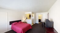Located at 2630 W Lincoln Ave in Anaheim, California, Room 105 is a ground-floor studio unit offering a practical and self-sufficient layout. The main living area is open and carpeted, centered around a full-size bed with a nightstand and lamp. A wall-mounted closet door and a television atop a multi-drawer dresser add functional furnishings, while a small dining table with chairs provides a dedicated spot for meals or work. Natural light enters through a front-facing window that looks out to the exterior grounds. A half-wall partition separates the kitchenette from the sleeping area, creating a defined cooking space equipped with dark wood cabinetry, a microwave, and a full-size refrigerator. The tiled kitchenette floor contrasts with the dark carpeting throughout the rest of the room. A separate vanity area features a sink with a light countertop and dark cabinetry, positioned adjacent to an open closet alcove with a full-width hanging rod and shelf — offering ample storage for extended stays. The bathroom is accessible from the vanity area. With a textured white ceiling throughout, the room presents a straightforward and functional design suited for short- or long-term occupancy in the heart of Anaheim.