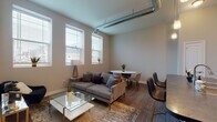 Located at 751 E Wheeling St in Lancaster, Ohio, this single-floor unit blends contemporary design with industrial-inspired details to create a functional and stylish living space. The open-concept living and dining area is immediately notable for its tall ceilings and exposed metallic ductwork, lending the space an urban loft aesthetic. Large, vertically oriented windows line the walls, drawing in abundant natural light and offering views of the surrounding neighborhood. Wood-look plank flooring runs throughout, providing a warm, cohesive foundation across all rooms.

The kitchen features white cabinetry extending to the upper walls, dark countertops, and a peninsula that provides additional workspace and casual seating. Black appliances, including a range, microwave, and refrigerator, complement the kitchen's modern palette.

The bedroom is a well-proportioned retreat with tall ceilings, large windows for natural light, and continued wood-look flooring. Exposed ductwork carries the industrial design theme into this space as well. The full bathroom is finished with white cabinetry, dark countertops, and a tub-shower combination. Throughout, neutral wall tones create a clean, modern ambiance suited to a range of furnishing styles.