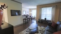 This single-story space showcases an open-concept floor plan that seamlessly connects the living, dining, and bar areas into one cohesive environment. The living area is anchored by a wall-mounted flat-screen television and features a patterned area rug atop dark hardwood-style flooring, which extends throughout the space, adding warmth and continuity. A stylish oval coffee table serves as a central focal point in the sitting area.

A built-in bar counter with tall barstool seating creates a natural transition between the living and dining areas, offering a casual spot for entertaining or everyday use. Beyond the bar, a dedicated dining area accommodates a table with several chairs beneath a pendant light fixture, adding an elegant touch overhead.

Large windows dressed with horizontal blinds and floor-length draping panels allow generous natural light to fill the space, creating a bright and welcoming atmosphere. Crown molding details along the upper walls lend a refined, polished character to the interior. Neutral wall tones and warm lighting contribute to an overall ambiance that feels both comfortable and tastefully appointed.