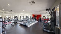 This fitness center offers a comprehensive workout environment spread across a single floor, thoughtfully organized into distinct exercise zones. The space is characterized by its dark rubber flooring, recessed lighting, and large arched and rectangular windows that flood the interior with natural light while providing views of the surrounding greenery.

The cardio zone is prominently featured, housing multiple Matrix-brand treadmills, elliptical trainers, and stair climbers arranged in orderly rows. A bold red accent wall serves as a visual anchor at one end of the space, complemented by wall-mounted televisions for entertainment during workouts. Large mirrors are strategically placed along the walls, enhancing the sense of openness throughout.

The strength training area includes a multi-functional cable machine, a free-weight station with a full rack of dumbbells and barboyts, adjustable benches, and a selection of kettlebells and medicine balls. A dedicated bay with expansive windows houses additional cable and weight equipment, benefiting from abundant natural light and outdoor views.

The facility's neutral-toned walls, accented by the striking red feature wall, create an energizing atmosphere. The overall layout is open and well-organized, providing ample space for a wide range of fitness activities.