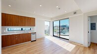 Located on Santa Monica Boulevard in the heart of West Los Angeles, this modern one-bedroom, one-bathroom residence offers a thoughtfully designed open-concept layout. Light oak-toned hardwood flooring runs throughout, complementing the crisp white walls and recessed lighting that give the space a clean, contemporary feel.

The combined kitchen and living area forms the heart of the unit. The kitchen is outfitted with warm walnut-toned flat-front cabinetry extending to the ceiling, white quartz countertops, and a full suite of stainless steel appliances including a refrigerator, electric range, built-in microwave, and dishwasher. A narrow horizontal window above the sink brings in additional natural light. Large floor-to-ceiling sliding glass doors flood the living area with sunlight and open directly onto a private balcony featuring glass panel railings, offering open views of the surrounding Los Angeles cityscape and lush street-level greenery.

The bedroom is a comfortable and well-proportioned retreat, featuring a large picture window with city views and a mirrored sliding-door closet. Additional conveniences include an in-unit laundry area and a full bathroom. The clean architectural lines, abundant natural light, and private outdoor space make this residence a polished urban retreat in a prime Los Angeles location.