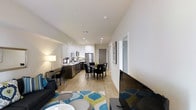 This single-floor space is designed around an open-concept layout that seamlessly connects the living, dining, and kitchen areas into one cohesive environment. Light hardwood flooring runs throughout, lending warmth and visual continuity across the space.

The living area is anchored by a large flat-screen television and a comfortable sofa, complemented by a vibrant area rug featuring teal, green, and white tones that adds a pop of color. A floor lamp provides ambient lighting alongside recessed ceiling lights that illuminate the entire space efficiently.

The dining area sits at the center of the unit, furnished with a dark rectangular table and matching chairs, offering a functional space for everyday meals. Adjacent to it, the kitchen features white shaker-style cabinetry, stainless steel appliances including a refrigerator and range, and a tile backsplash. The countertops provide ample workspace, and the layout supports ease of movement.

A hallway leading toward the rear of the unit suggests additional rooms beyond, with framed wall art adding a refined touch. The overall ambiance is bright, clean, and contemporary, with a neutral white palette that enhances the sense of openness throughout.
