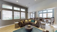 The conference room at 1100 Garden View in Encinitas is a thoughtfully designed, single-floor space that seamlessly blends comfort and functionality. Soaring ceilings create an open, airy atmosphere, while an abundance of large windows fitted with slatted blinds flood the interior with natural light and offer views of the surrounding greenery.

The lounge area serves as the centerpiece of the space, anchored by a generous L-shaped sectional sofa in warm charcoal upholstery, accented with teal, yellow, and neutral throw pillows. A round, sculptural coffee table sits atop a patterned teal area rug, complemented by a pair of decorative metallic side tables. The light hardwood flooring adds warmth and continuity throughout.

Beyond the lounge, the space transitions into a dining and work area furnished with tables and upholstered chairs, suitable for collaborative meetings or individual work sessions. The two-tone wall treatment, combining crisp white with deep taupe-gray, adds visual depth and a polished, contemporary aesthetic. Overall, the space offers a welcoming and professional environment well-suited for both meetings and casual gatherings.