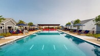 Located at 2370 East Centerville Road in Garland, Texas, this single-level outdoor amenity space is designed to deliver a resort-style experience for its residents. At the heart of the area is a large, rectangular swimming pool with crystal-clear blue water, framed by a spacious concrete deck that provides ample room for relaxation and socializing.

The pool deck is thoroughly appointed with a generous collection of cushioned lounge chairs in bold navy and red, accompanied by striped navy-and-white market umbrellas that provide shade throughout the day. The vibrant color scheme gives the space an energetic yet welcoming ambiance.

A covered pergola structure anchors one end of the deck, featuring a warm wood-paneled ceiling supported by steel columns. This shaded area includes seating and offers views of the surrounding neighborhood, making it an ideal gathering spot during warmer hours. Additional open-air seating with red chairs and white tables is also available for casual outdoor dining or conversation.

The pool area is enclosed by black iron fencing with gated access, providing both security and a clean aesthetic that complements the community's architectural style. The surrounding buildings feature white brick, board-and-batten siding, and pitched gray rooflines, creating a cohesive, contemporary neighborhood feel. Landscaping with ornamental grasses and young trees adds a natural touch to the overall setting.