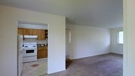 Located at 25 Bishop Terrace in Stratford, New Jersey, this single-floor home presents a practical and functional layout. The living area features neutral-toned carpeting that extends throughout, creating a cohesive and understated aesthetic. The space is defined by clean white walls and standard ceiling heights, contributing to a bright and open atmosphere.

Adjacent to the living area, the kitchen is accessible through an open pass-through design, allowing for a connected flow between the two spaces. The kitchen is outfitted with warm wood-toned cabinetry offering ample storage, tile backsplash detailing, and tile flooring. A white freestanding range serves as the primary cooking appliance, complementing the classic, utilitarian character of the kitchen.

A doorway visible from the main living space suggests additional rooms beyond, providing further separation within the home's layout. The overall design of this single-floor residence prioritizes functionality, with straightforward finishes and an efficient use of space throughout.