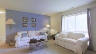 This single-floor space centers around a well-appointed living room that combines comfort and style through a carefully chosen color scheme. A striking periwinkle-blue accent wall serves as a focal point, contrasting elegantly with the surrounding soft cream-toned walls and light-colored carpeted flooring. The room accommodates two plush white slipcovered sofas arranged around a decorative wrought-iron and wood coffee table, creating a cozy and functional seating area. Accent pillows in chevron and geometric patterns add visual interest to the neutral upholstery. A large window dressed with white blinds and sheer curtains allows natural light to filter into the space, with views of greenery visible outside. Warm ambient lighting is provided by a classic table lamp, complementing the natural light throughout the day. The accent wall is adorned with four framed artworks arranged in a symmetrical grid pattern, adding a personal and polished touch. Decorative elements such as a floor vase with branches and potted greenery enhance the organic, serene ambiance of the room.