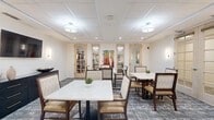 This thoughtfully designed communal room offers a refined and comfortable setting for dining or group gatherings. The space accommodates multiple square tables, each topped with white surfaces and surrounded by upholstered arm chairs featuring a distinctive geometric pattern and warm tan leather seat cushions — providing both style and comfort for occupants.

The room is finished with a neutral color palette of soft whites and warm beiges, complemented by a decorative patterned carpet in grey and white tones that adds visual depth to the floor. Overhead, a drop tile ceiling is fitted with a combination of flush-mount drum pendant lights and recessed lighting, ensuring even, well-distributed illumination throughout.

A wall-mounted flat-screen television is positioned on one wall alongside a dark credenza, offering both functional media access and additional storage. Wall sconces provide supplemental ambient lighting on either side of the room. A vibrant, colorful abstract painting serves as a focal art piece, adding a lively contrast to the otherwise subdued decor.

Multiple French-style glass-panel doors with light wood frames line the perimeter, connecting this room to adjacent spaces — including what appears to be a library and a lounge area — allowing for natural light flow and flexible use of the surrounding facility. Small potted plants at each table add a fresh, welcoming touch.
