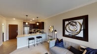This thoughtfully designed single-floor residence offers a seamless open-concept layout connecting the kitchen and living area. The kitchen is a standout feature, showcasing rich dark-wood cabinetry, granite countertops, a peninsula with bar seating illuminated by pendant lighting, and a full suite of stainless steel appliances including a refrigerator, electric range, microwave, and dishwasher. Wood-look flooring runs throughout the kitchen, transitioning to plush carpet in the living and sleeping areas. Crown molding adds a refined architectural detail throughout the main living spaces. The living area is bright and comfortable, with access to a balcony that brings in natural light and outdoor views. The bedroom is generously proportioned, featuring a large window, ample closet space, and direct access to a walk-in closet that also houses a stacked Whirlpool washer and dryer — a highly practical in-unit convenience. The full bathroom is cleanly finished with dark granite countertops, a rich wood vanity, a tub-shower combination, and wood-look flooring that ties the space together. Neutral warm tones and consistent finishes throughout create a cohesive, modern ambiance.