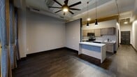 Located at 229 W Third St in downtown Davenport, Iowa, this one-bedroom, one-bathroom unit offers a well-considered layout across a single floor. The open-concept living and kitchen area is anchored by a striking marble-topped island with pendant lighting overhead, complemented by gray cabinetry, stainless steel appliances, and rich dark hardwood flooring that runs throughout the main living space. An oversized ceiling fan adds both function and a modern industrial accent to the room. A bank of tall windows floods the living area with natural light and frames urban views of the surrounding cityscape, with curtain panels adding a warm, residential touch. The bedroom features plush carpeting, bold black-trimmed doors, a closet, and elevated ceilings that enhance the sense of space. A ceiling fan continues the consistent design language found throughout the unit. The bathroom is thoughtfully appointed with a wood-toned vanity, white countertop, round mirrors with matte black hardware, and warm wall sconces. Conveniently integrated into the bathroom area is a stacked washer and dryer unit, providing in-unit laundry without sacrificing additional living space. A connecting hallway ties all areas together cohesively, making this a practical and stylish urban residence.