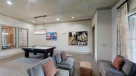 This single-floor amenity space is thoughtfully designed to offer a welcoming and versatile social environment. The game room serves as a central highlight, featuring a full-size pool table anchored beneath a modern rectangular pendant light fixture that adds a sophisticated focal point to the room. A wall-mounted flat-screen television is positioned nearby, making the space ideal for both gaming and entertainment.

The seating area is furnished with plush, generously sized gray sofas and accent chairs adorned with warm-toned throw pillows, creating a comfortable and inviting lounge atmosphere. A leather ottoman adds a contrasting texture to the arrangement. The flooring consists of wood-style planks in a neutral tone that complement the light, neutral-painted walls throughout.

Recessed lighting provides consistent, ambient illumination across the ceiling, which features subtle crown molding detail for an added architectural touch. The walls are decorated with striking black-and-white photographic art prints depicting urban scenes, lending the space a contemporary and cosmopolitan character. An adjacent doorway suggests additional rooms or access points beyond the main lounge area. Overall, the space balances functionality with modern aesthetic appeal.