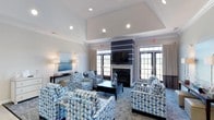 Located at 1010 Fairway Village in Whitsett, North Carolina, this single-floor community space offers a welcoming and well-appointed lounge area designed for comfort and social gathering. The room's centerpiece is a classic fireplace framed by a bold navy blue accent wall, above which a flat-screen television is mounted, creating a functional yet elegant focal point. The seating area is furnished with a coordinated arrangement of patterned blue and white geometric armchairs surrounding a dark coffee table, providing an inviting atmosphere for relaxation or conversation. An elevated tray ceiling with recessed lighting adds a sense of height and architectural refinement to the space. Large French-style doors and surrounding transom windows line the walls, flooding the interior with abundant natural light while offering views of the surrounding outdoor environment. Neutral wall tones and cream drapery complement the coastal-inspired décor, including ocean-themed artwork that reinforces a calm and serene ambiance. The combination of thoughtful design details, quality furnishings, and generous natural light makes this community lounge a comfortable and attractive gathering place for residents of Fairway Village.