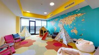 Located at 1631 Paterson Plank Rd in Secaucus, NJ, this single-floor children's playroom is designed to engage and inspire young minds through bold, stimulating design. The space immediately draws attention with its vivid yellow ceiling accents contrasting against cream-toned walls, creating an energetic yet welcoming atmosphere throughout.

The most striking feature is a large-scale illustrated world map mural covering an entire teal-painted accent wall, depicting continents, oceans, and illustrated wildlife in a colorful, educational style. This serves as both a decorative centerpiece and an interactive learning element for children.

The flooring features a dynamic arrangement of hexagonal carpet tiles in shades of red, orange, yellow, green, and gray, adding visual interest and defining different activity zones within the room. Play structures include decorative teepees, a wooden playhouse, and a child's slide, offering imaginative play opportunities.

A designated lounge area provides comfortable seating for adults and older children, furnished with bean bag chairs, a pink woven accent chair, and a small wooden side table. A wall-mounted flat-screen television and a row of animal-themed storage cubbies complete the space. Large windows and glass doors along one wall invite natural light and provide views of an outdoor area.
