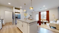This single-floor residence is thoughtfully designed with a contemporary aesthetic and an efficient, open layout. The kitchen serves as a central highlight, featuring white shaker cabinetry with gold-toned hardware, granite countertops, a generously sized center island with bar seating, and a full suite of stainless steel appliances. Two globe pendant lights with brass accents hang above the island, complementing the recessed lighting and light wood-look flooring that runs throughout the space.

The kitchen flows seamlessly into the living area, which benefits from natural light through large windows dressed with horizontal blinds and warm rust-colored drapes. A glass door provides additional light and outdoor access. A ceiling fan adds comfort to the living space, which features a slate-blue accent wall.

The bedroom is a composed retreat with a deep plum accent wall, natural light from a large window with sheer curtains, and an adjacent full bathroom. The bathroom features granite-topped white cabinetry, a tiled tub-shower combination, and light wood-look flooring. A spacious walk-in closet with double hanging rods provides ample storage.