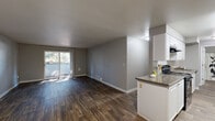 Located at 4820 NE Hazel Dell Ave in Vancouver, Washington, this one-bedroom, one-bathroom residence offers a well-organized single-floor layout with a comfortable sense of space throughout. The combined kitchen and living area forms the heart of the home, with the living room showcasing rich, dark hardwood-style flooring, neutral gray walls, and a sliding glass door that opens to a private balcony overlooking lush greenery — allowing natural light to fill the space. A baseboard heating unit ensures year-round comfort. The galley-style kitchen is efficiently designed with white cabinetry lining both walls, granite-toned countertops, wood-look flooring, and black appliances including a range, dishwasher, and refrigerator, providing ample storage and functionality. The bedroom features plush carpeting, a bright window with outdoor views, a baseboard heater, and a clean, neutral palette that creates a calm retreat. The full bathroom is finished with wood-look flooring, white cabinetry, a tiled shower-tub combo, and tasteful neutral tones that tie the space together cohesively. Overall, the unit presents a clean, modern aesthetic with practical finishes suited for comfortable everyday living.