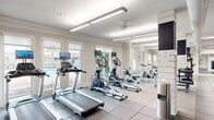This fitness center in Cayce, South Carolina, offers a comprehensive and thoughtfully organized workout facility across a single floor. The space is divided into two distinct zones: a bright cardio area and a dedicated strength training section, providing a well-rounded environment for various fitness routines.

The cardio zone is lined with large windows along multiple walls, flooding the area with natural light and offering views of the surrounding outdoor pool area and landscaping. Several treadmills, elliptical machines, stationary bikes, and a rowing machine are arranged in rows, each equipped with individual screens for entertainment during workouts. Durable rubber-tiled flooring runs throughout, providing both safety and functionality.

The strength training area features a sleek, modern aesthetic with navy accent walls and large wall mirrors that amplify the sense of space. This section is equipped with adjustable weight benches, a dumbbell rack, cable machines, and additional strength equipment. The mirrored walls enhance visibility and create an open, expansive feel within the room.

Overhead, long linear pendant lighting and flush-mounted fixtures provide consistent, even illumination throughout. The neutral color palette of white and gray walls, accented by dark tones, contributes to a clean and motivating atmosphere. Motivational signage adds an encouraging touch, completing this well-appointed fitness facility.