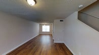 This two-floor space presents a straightforward and functional layout with notable design details throughout. The main living area showcases rich hardwood flooring that extends seamlessly across the room, lending warmth and continuity to the space. Crisp white walls provide a neutral backdrop, complemented by white baseboards that frame the flooring neatly. The textured ceiling adds visual interest overhead, while a flush-mount light fixture provides overhead illumination. A pendant light is visible in the adjoining area, hinting at a defined transition between spaces. A window at the far end allows natural light to filter into the interior, brightening the open floor plan. A wall-mounted HVAC vent is discreetly positioned along the upper portion of the wall, ensuring climate control throughout. The overall ambiance is clean, open, and move-in ready, with a simple yet inviting character. The two-floor layout offers additional living space beyond what is visible, providing a sense of depth and versatility to the overall floor plan.