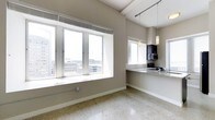 This contemporary single-floor space offers a bright, open layout designed to maximize both functionality and visual appeal. The living area is anchored by a generous bank of large windows that frame expansive cityscape views, flooding the interior with natural light and creating a strong connection to the urban environment outside. The neutral, warm-gray wall palette and polished terrazzo-style flooring work together to give the space a clean, sophisticated character throughout.

The kitchen is seamlessly integrated into the open floor plan, featuring dark cabinetry, granite countertops, and a practical peninsula that serves as both a workspace and a casual dining surface. A pendant light fixture provides focused illumination over the counter, complementing the track lighting installed along the ceiling beams. A stainless steel microwave and sink are neatly incorporated into the cabinetry, ensuring the space is as functional as it is visually cohesive.

Architectural details such as the exposed ceiling beams and generous ceiling height add a sense of volume and industrial character to the space. The combination of abundant natural light, polished surfaces, and panoramic city views makes this a compelling and well-appointed urban space.