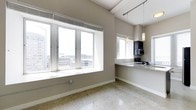 This contemporary single-floor space offers a bright, open layout designed to maximize both functionality and visual appeal. The living area is anchored by a generous bank of large windows that frame expansive cityscape views, flooding the interior with natural light and creating a strong connection to the urban environment outside. The neutral, warm-gray wall palette and polished terrazzo-style flooring work together to give the space a clean, sophisticated character throughout.

The kitchen is seamlessly integrated into the open floor plan, featuring dark cabinetry, granite countertops, and a practical peninsula that serves as both a workspace and a casual dining surface. A pendant light fixture provides focused illumination over the counter, complementing the track lighting installed along the ceiling beams. A stainless steel microwave and sink are neatly incorporated into the cabinetry, ensuring the space is as functional as it is visually cohesive.

Architectural details such as the exposed ceiling beams and generous ceiling height add a sense of volume and industrial character to the space. The combination of abundant natural light, polished surfaces, and panoramic city views makes this a compelling and well-appointed urban space.