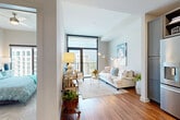 This thoughtfully designed single-floor residence offers a seamless blend of modern style and everyday functionality. The open-concept living area is anchored by rich hardwood flooring and flooded with natural light through floor-to-ceiling windows and a glass balcony door, offering views of the surrounding cityscape. The living room provides a welcoming, airy atmosphere, complemented by neutral tones and curated artistic touches.

The kitchen is a standout feature, showcasing gray shaker-style cabinetry, open shelving, a white countertop island, stainless steel appliances, and striking gold-accented pendant lighting — all set atop warm hardwood floors. The layout is both practical and elegant, ideal for cooking and entertaining.

The bedroom is a calm retreat, carpeted for comfort, with a large window that frames outdoor views and fills the room with natural light. The bathroom is particularly eye-catching, featuring bold black-and-white patterned floor tiles, gray cabinetry with gold hardware, a white quartz vanity, and a conveniently integrated stacked washer and dryer unit. Throughout the space, a cohesive palette of warm neutrals, metallic accents, and natural materials creates a polished, contemporary ambiance.
