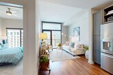 This thoughtfully designed single-floor residence offers a seamless blend of modern style and everyday functionality. The open-concept living area is anchored by rich hardwood flooring and flooded with natural light through floor-to-ceiling windows and a glass balcony door, offering views of the surrounding cityscape. The living room provides a welcoming, airy atmosphere, complemented by neutral tones and curated artistic touches.

The kitchen is a standout feature, showcasing gray shaker-style cabinetry, open shelving, a white countertop island, stainless steel appliances, and striking gold-accented pendant lighting — all set atop warm hardwood floors. The layout is both practical and elegant, ideal for cooking and entertaining.

The bedroom is a calm retreat, carpeted for comfort, with a large window that frames outdoor views and fills the room with natural light. The bathroom is particularly eye-catching, featuring bold black-and-white patterned floor tiles, gray cabinetry with gold hardware, a white quartz vanity, and a conveniently integrated stacked washer and dryer unit. Throughout the space, a cohesive palette of warm neutrals, metallic accents, and natural materials creates a polished, contemporary ambiance.