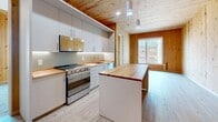 Located at 29 Crown Street in New Haven, CT, this 2-bedroom, 2-bathroom unit showcases a striking architectural identity rooted in mass timber construction. Exposed knotty pine panels line the walls and ceilings throughout, imparting a warm, natural aesthetic that contrasts beautifully with modern finishes. The open-plan kitchen and living area serves as the heart of the unit. The kitchen is outfitted with sleek gray cabinetry extending to the ceiling, stainless steel appliances including a full-range stove and dishwasher, a single-basin sink, and rich butcher block countertops. A central island with a matching butcher block surface provides additional prep and dining space, with pendant lighting overhead. Light wood-look flooring runs throughout the main living areas, complementing the natural pine elements. Both bedrooms feature soft carpet flooring and white walls offset by the signature pine ceiling. The primary bedroom includes built-in white cabinetry for storage. Large windows in the bedrooms invite natural light and offer urban views of the surrounding New Haven cityscape. The two bathrooms are finished in crisp white, with wood-look tile flooring — one featuring a walk-in shower and the other a full soaking tub. An in-unit laundry area and a dedicated entrance complete this thoughtfully designed urban residence.