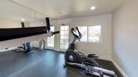 Located at 1539 Tavern Road in Alpine, California, this dedicated fitness facility offers a well-equipped, single-floor workout space designed to support a well-rounded exercise routine. The room features durable dark rubber flooring that provides both safety and stability during training sessions, along with recessed lighting and bright white walls that create a clean, functional atmosphere.

The space is outfitted with a multi-station strength training system — a Body-Solid unit — providing a range of resistance-based exercises including cable pulldowns, chest press, and leg workouts. Free weight plates are also available for added versatility. Complementing the strength equipment is an elliptical cross-trainer, ideal for low-impact cardiovascular exercise.

Full-length mirrors line one wall, enhancing the sense of space and allowing users to monitor their form during workouts. Multiple windows frame views of the surrounding outdoor greenery, filling the room with natural light and offering pleasant outdoor sightlines. A glass-paned door provides direct exterior access, further connecting the interior to the natural surroundings.

Posted exercise rules and additional informational signage reflect an organized, community-oriented environment. The overall layout is efficient and practical, making this fitness space a functional amenity for those seeking a convenient and well-maintained place to exercise.