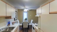 This single-floor residence offers a thoughtfully arranged layout that combines everyday functionality with a warm, inviting ambiance. The kitchen and dining area are seamlessly integrated, creating an open and efficient use of space. The kitchen is outfitted with white cabinetry that extends to the upper walls, providing ample storage, while white appliances — including an electric range and refrigerator — offer practical cooking amenities. Light countertops and a sink complete the workspace. Adjacent to the kitchen, the dining area features a round glass-top table surrounded by upholstered white chairs, perfectly suited for intimate meals. Warm earth-tone walls give the space a cozy, grounded feel, while dark hardwood-style flooring adds a rich contrast throughout. A ceiling fan with an integrated light fixture serves as a central focal point, ensuring comfortable airflow and ambient lighting. A window dressed with horizontal blinds allows natural light to filter into the dining area, brightening the space. Decorative touches, including a framed wall print and a small potted plant, add personality and a lived-in warmth to the overall environment.