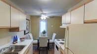 This single-floor residence offers a thoughtfully arranged layout that combines everyday functionality with a warm, inviting ambiance. The kitchen and dining area are seamlessly integrated, creating an open and efficient use of space. The kitchen is outfitted with white cabinetry that extends to the upper walls, providing ample storage, while white appliances — including an electric range and refrigerator — offer practical cooking amenities. Light countertops and a sink complete the workspace. Adjacent to the kitchen, the dining area features a round glass-top table surrounded by upholstered white chairs, perfectly suited for intimate meals. Warm earth-tone walls give the space a cozy, grounded feel, while dark hardwood-style flooring adds a rich contrast throughout. A ceiling fan with an integrated light fixture serves as a central focal point, ensuring comfortable airflow and ambient lighting. A window dressed with horizontal blinds allows natural light to filter into the dining area, brightening the space. Decorative touches, including a framed wall print and a small potted plant, add personality and a lived-in warmth to the overall environment.