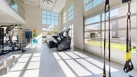 Located at 8901 Holly Ave NE in Albuquerque, New Mexico, this fitness center offers a bright, modern exercise space designed to support a full range of workout routines. The room features dramatically high ceilings with expansive floor-to-ceiling windows and large grid-pane glass panels on multiple walls, flooding the space with natural light and providing open views of the surrounding neighborhood and landscaping. A glass-railed upper mezzanine element and recessed lighting contribute to the airy, contemporary feel of the interior. The light wood-toned flooring and crisp white walls create a clean, energizing atmosphere throughout.

The space is comprehensively equipped for both strength training and cardiovascular exercise. Strength training amenities include a multi-station cable machine, adjustable weight benches, a rack of dumbbells, and a dedicated TRX suspension training station with accessories. Cardio equipment includes treadmills, elliptical machines, and a stair climber, all positioned to take advantage of the natural light and outdoor views. A selection of stability and medicine balls rounds out the functional fitness offerings. Full-length mirrors along one wall enhance the sense of openness while providing a practical training aid. A ceiling fan supports ventilation and comfort during workouts.