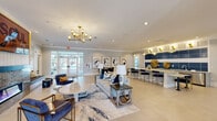 Located at 4692 E Ponce de Leon Ave in Stone Mountain, Georgia, this single-story community clubhouse offers a thoughtfully designed gathering space with a cohesive modern aesthetic. The entry opens into a grand vaulted foyer framed by white brick columns, setting an elegant tone before flowing into the main lounge area. The central lounge is anchored by a striking wood-surround fireplace with a stone hearth, accompanied by curated seating arrangements, a geometric gold wall installation, and a built-in bench nook. A bold gold chandelier adds warmth and sophistication overhead. Adjacent to the lounge, a stylish wet bar features a large granite-topped island with navy velvet barstools, bold blue tile accents, and gold pendant lighting, creating a vibrant social hub. Multiple dining vignettes are scattered throughout, including a casual seating area with a wall-mounted TV and a Scrabble board, and a refined dining nook adorned with a series of large abstract artworks. A well-equipped kitchenette with wood cabinetry, granite countertops, and a coffee station supports the dining areas. The business center provides multiple computer workstations with ample natural light from surrounding windows. Light tile flooring, neutral gray walls, and curated artwork unify the space with a polished, contemporary character.