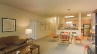 This single-floor residence is designed around an open-concept layout that seamlessly connects the living, dining, and kitchen areas into one cohesive space. The living area is anchored by a sofa and a low wooden coffee table, complemented by a classic table lamp that casts a warm, inviting glow. Neutral-toned carpet extends throughout, adding softness underfoot and contributing to the overall warmth of the interior.

The dining area features a glass-top table surrounded by rattan-framed chairs with red upholstered cushions, offering a casual yet refined setting for meals. A pendant chandelier with multiple globe lights hangs above, providing focused illumination over the dining space.

Adjacent to the dining area, the kitchen is equipped with light wood cabinetry, standard appliances including a microwave, and counter space visible from the open layout. A peninsula separates the kitchen from the dining area, enhancing both functionality and flow.

Along the walls, a series of framed botanical prints adds decorative interest, while a tall decorative branch arrangement near the kitchen contributes a natural element. The overall ambiance is warm, comfortable, and traditionally styled.