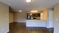 This single-floor residence offers a thoughtfully designed open-concept layout that seamlessly connects the kitchen and living areas, creating a spacious and welcoming atmosphere. The kitchen serves as a central focal point, showcasing dark espresso cabinetry that provides a striking contrast against the light granite countertops. Stainless steel appliances, including a refrigerator, range, and built-in microwave, lend a contemporary, professional feel to the space. A peninsula-style counter extends outward, offering additional prep space and the opportunity for casual seating, making it ideal for both cooking and entertaining. Pendant lighting hangs above the peninsula, adding a warm, stylish touch, while flush-mount ceiling fixtures illuminate the broader living area. Rich, dark hardwood-style flooring runs throughout the open space, adding warmth and visual continuity. The neutral, light-toned walls enhance the brightness of the interior, creating an airy and inviting ambiance. The living area provides generous room for a variety of furniture arrangements, and a nearby entry door adds convenient access. Overall, the space combines modern finishes with a functional, open design.