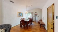 This single-floor space exudes a distinctive Spanish Colonial character, blending classic architectural details with warm, inviting interiors. Rich, dark hardwood floors run throughout, anchoring each room with a sense of depth and tradition. The main living area is a generously proportioned room, highlighted by striking teal-framed French doors that open directly onto a lush, plant-filled courtyard. A crystal chandelier adorns the ceiling, adding an elegant focal point, while built-in white shelving lines one wall, offering functional display space. Natural light streams in through the French doors, illuminating the open interior. A hallway with a dramatic barrel-vaulted ceiling connects the living areas, lending an airy, cathedral-like quality to the passage. This hall leads to additional rooms, including a bathroom that continues the Spanish aesthetic with terracotta tile floors, a classic soaking tub, white subway tile walls accented with decorative patterned inserts, and its own barrel-vaulted ceiling. The kitchen features white cabinetry, open shelving, wood countertops, and teal tile detailing around the sink that echoes the color palette found throughout. The private courtyard entrance, framed by climbing greenery and terracotta tile, completes the cohesive, character-rich design of this thoughtfully detailed space.